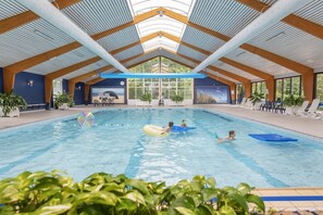 An indoor swimming pool with a high ceiling and wooden beams, surrounded by potted plants and seating areas. - Holiday Home Near Dutch Beach With Pool (Sint Maartensvlotbrug)