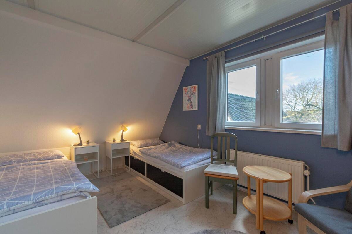 A small, well-lit room with two single beds, a chair, a small table, and a window providing a view of the outside.