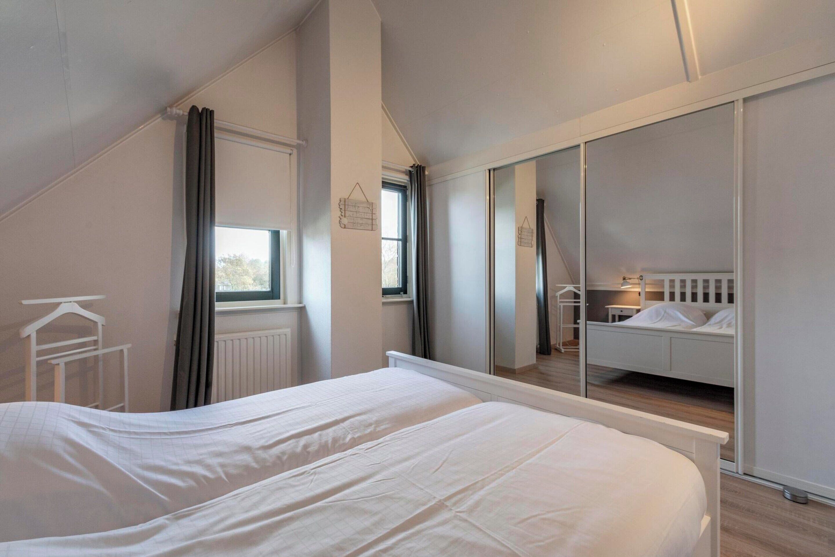 A bedroom with a large bed, a window with curtains, a radiator, and a mirror reflecting a bunk bed.