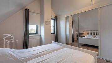 A bedroom with a large bed, a window with curtains, a radiator, and a mirror reflecting a bunk bed.