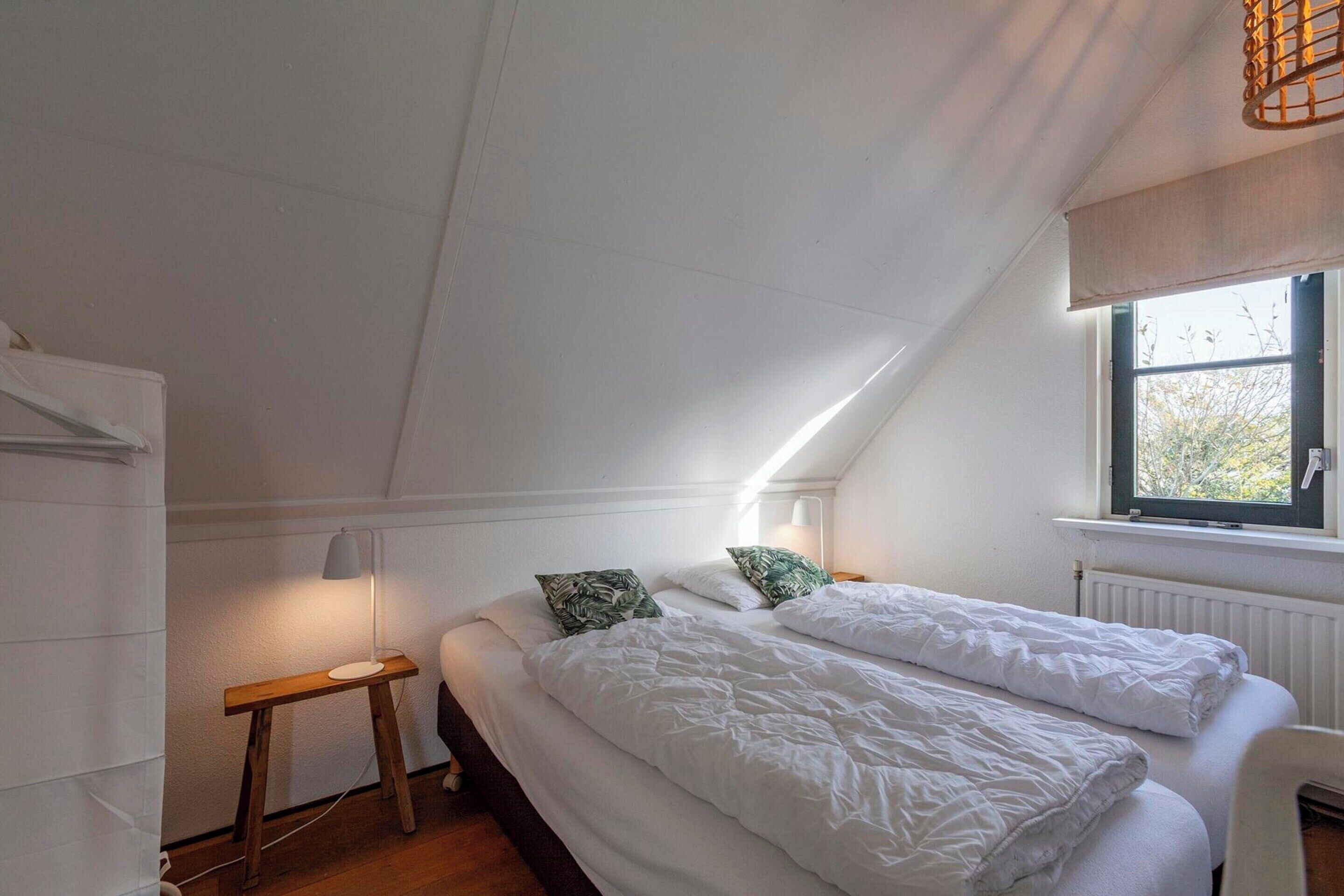Una camera da letto con un soffitto spiovente, una finestra vista alberi, un letto con biancheria bianca e un comodino con una lampada.