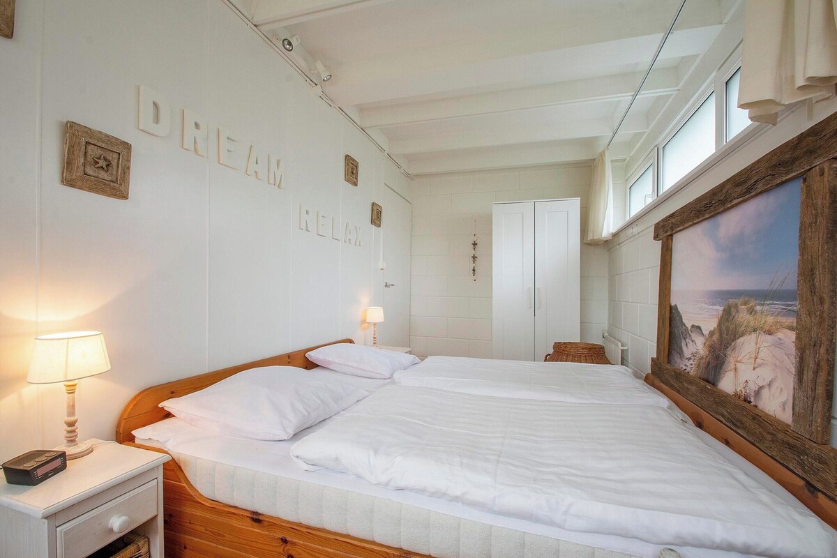 A bedroom with a large bed, two bedside tables, a nightstand, a wall-mounted lamp, and a framed painting.
