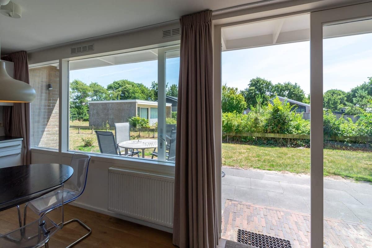 Une chambre moderne dotée d’un coin repas, de grandes fenêtres et offrant une vue sur un jardin et les maisons voisines.