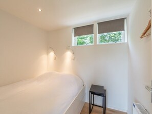 A bedroom with a bed, two bedside lamps, a small table, a window with blinds, and a radiator. - Bungalow in Callantsoog Near Sandy Beaches (Callantsoog)