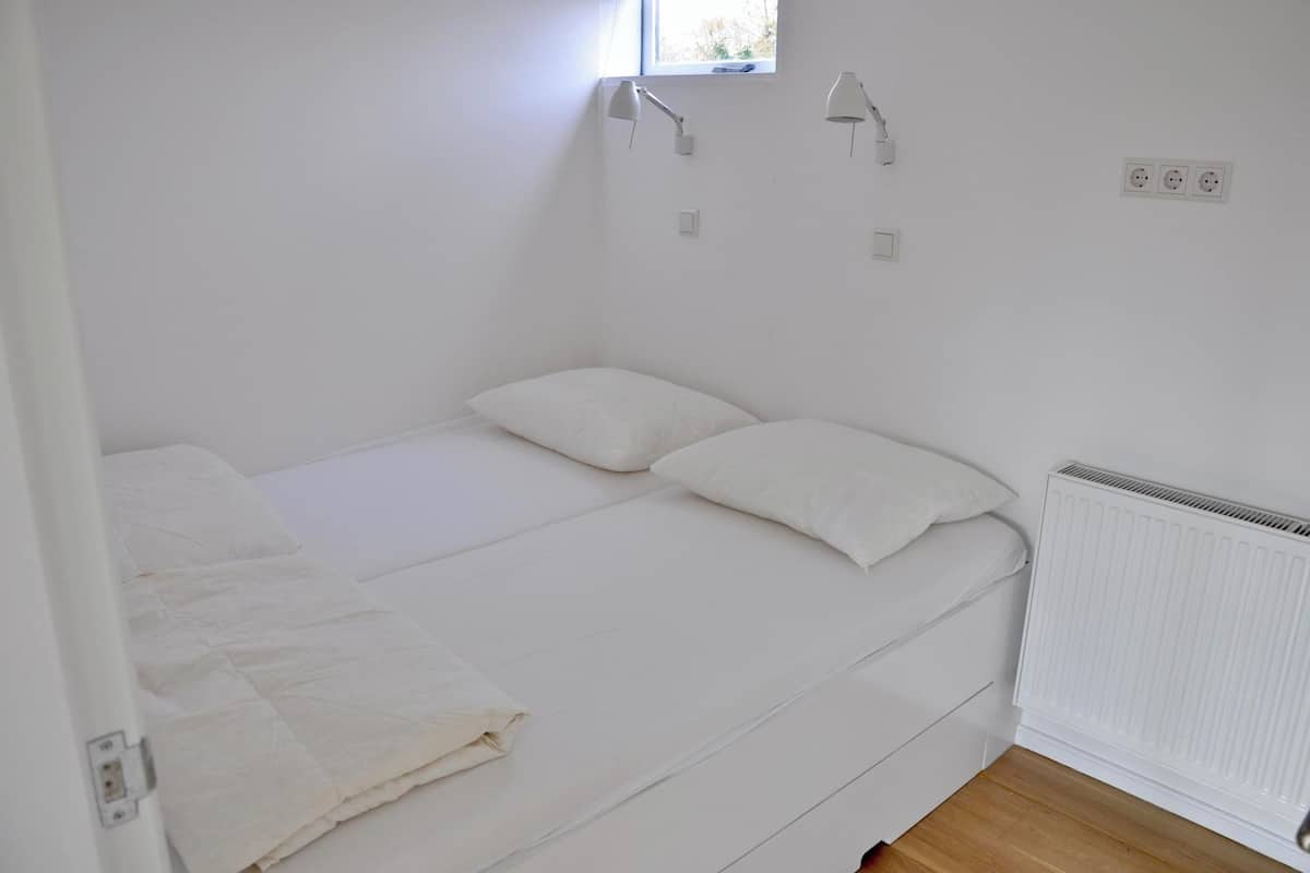 Une petite chambre avec un lit simple, une fenêtre, un radiateur et deux lampes fixées au mur.