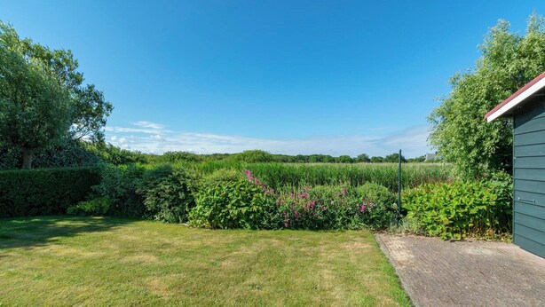 A grassy yard with a paved path, dense shrubbery, and a building with a red roof. - Villa in Callantsoog Near the Beach (Callantsoog)