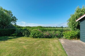 A grassy yard with a paved path, dense shrubbery, and a building with a red roof. - Villa in Callantsoog Near the Beach (Callantsoog)