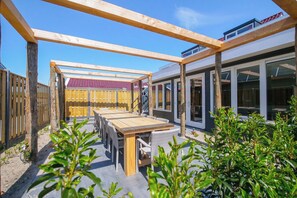 An outdoor patio with a wooden pergola, a dining table with chairs, and a view of a modern building with large windows. - Callantsoog Group Getaway Beach (Callantsoog)