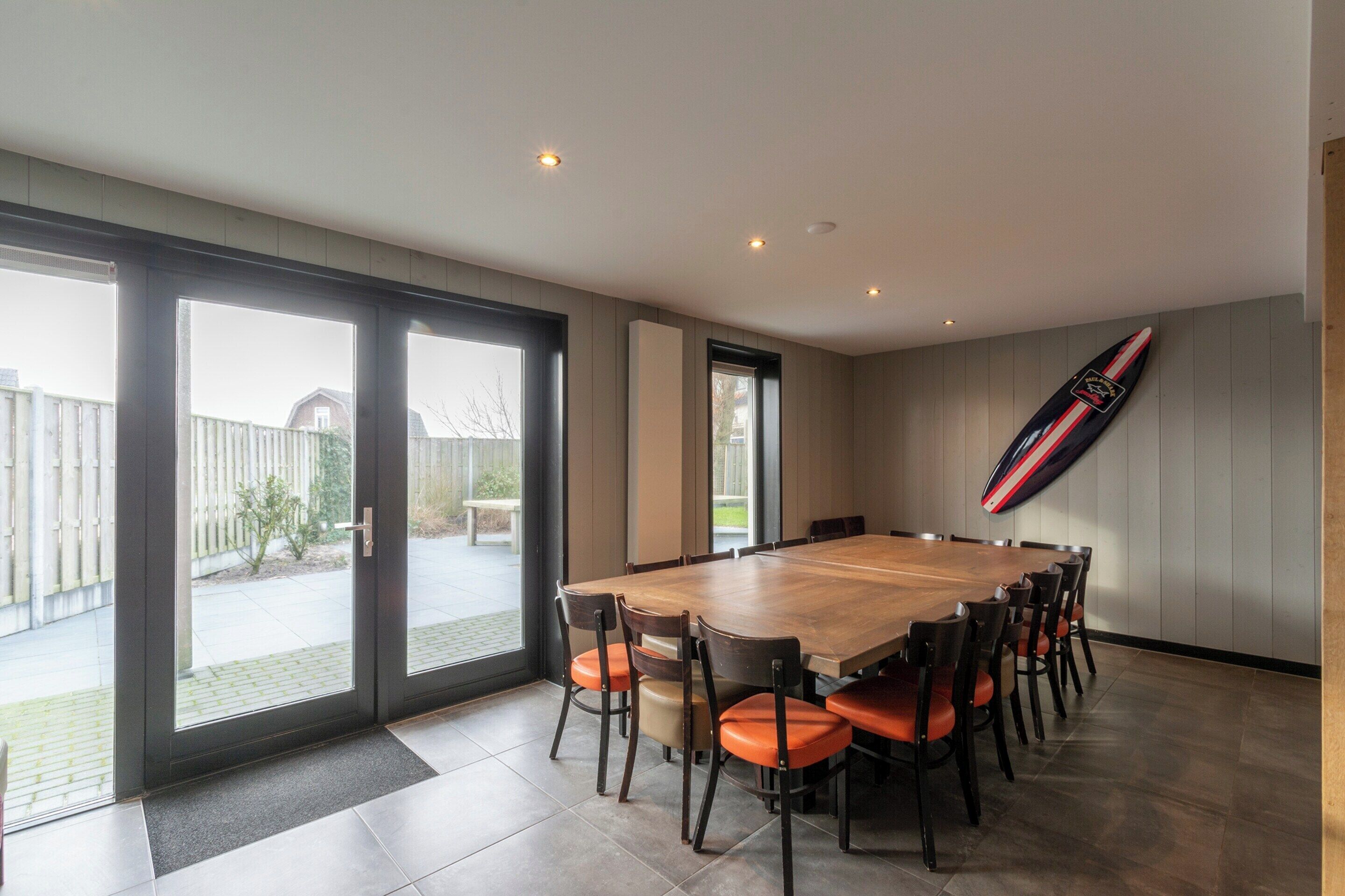 A spacious dining area with a large wooden table and chairs, large windows, and a surfboard on the wall.