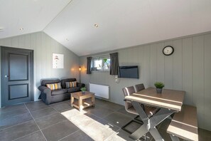Habitación con sofá, una mesita, una mesa de comedor con sillas, televisor montado en la pared y un reloj.
