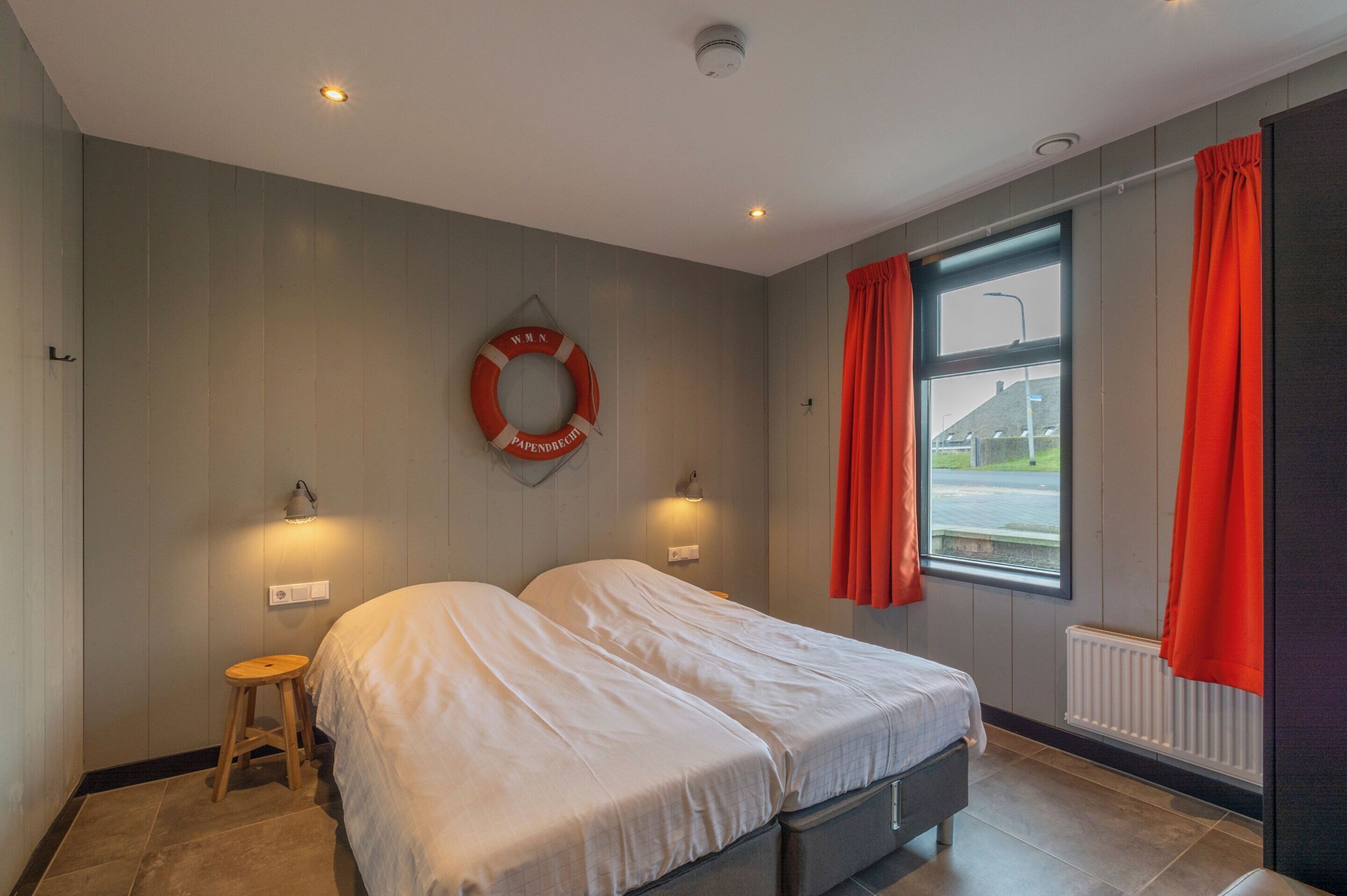 A bedroom with two beds, a small stool, a life preserver on the wall, and a window with curtains.