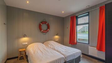 A bedroom with two beds, a small stool, a life preserver on the wall, and a window with curtains.