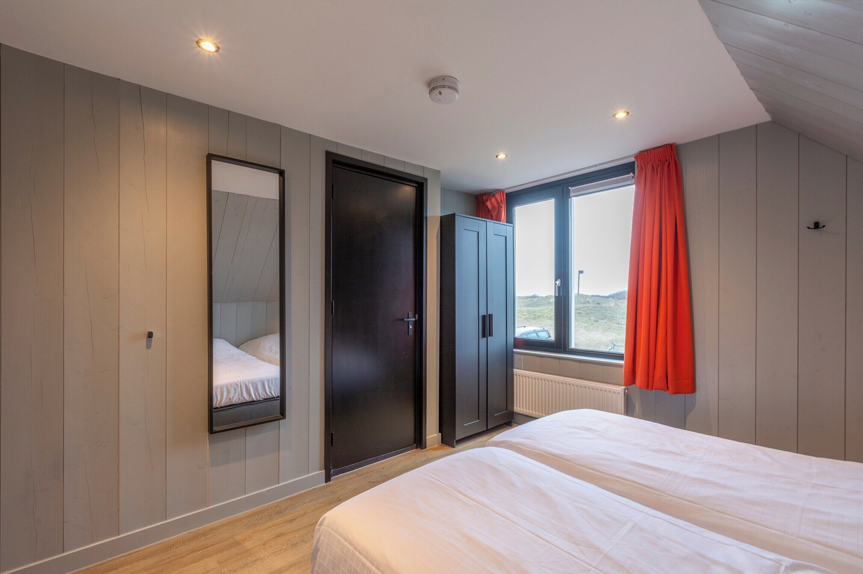 Un dormitorio con cama, armario, espejo y ventana con cortinas.