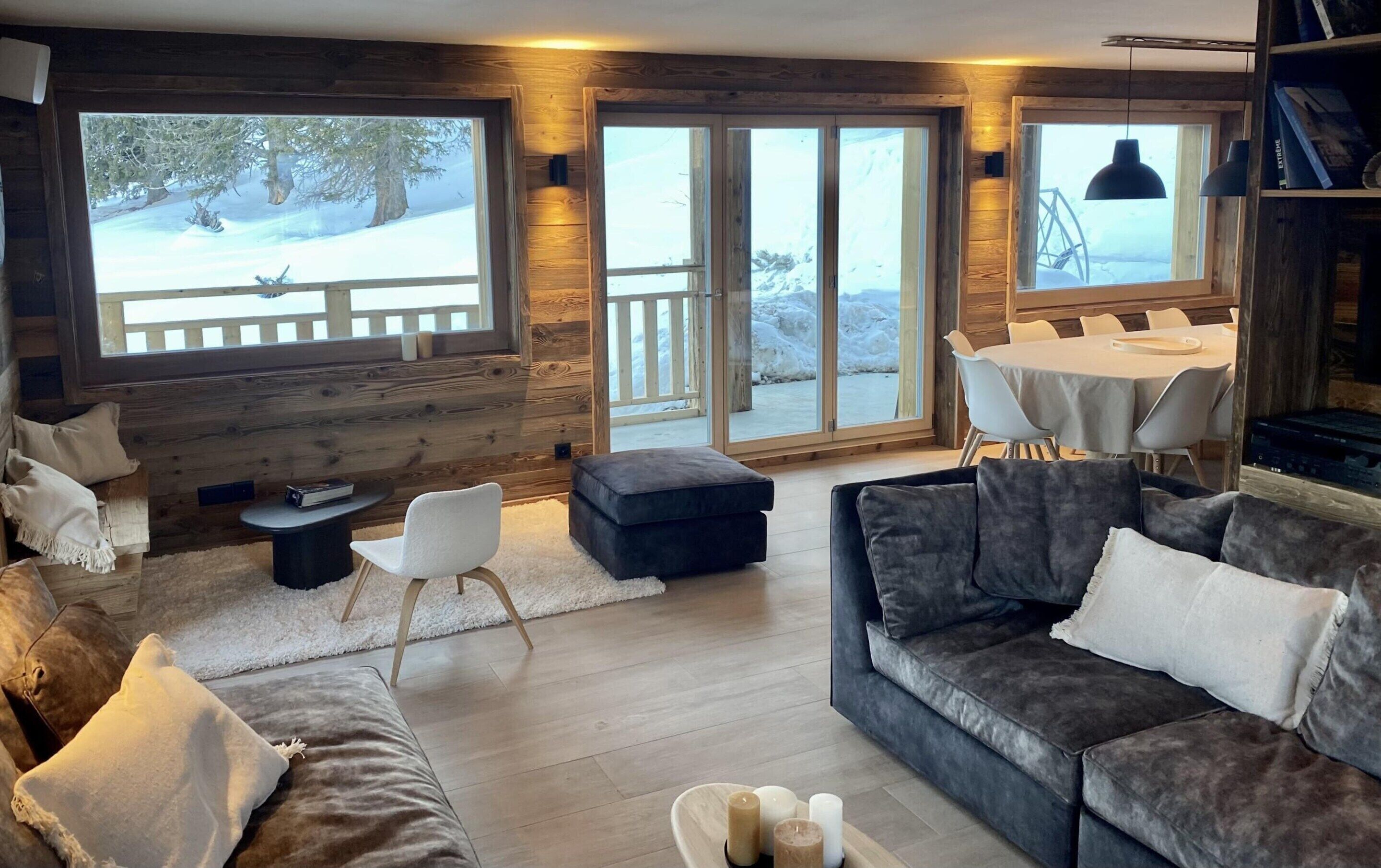 L’intérieur chaleureux d’un chalet avec une cheminée, des murs en bois, un coin repas et de grandes fenêtres donnant sur un paysage enneigé.