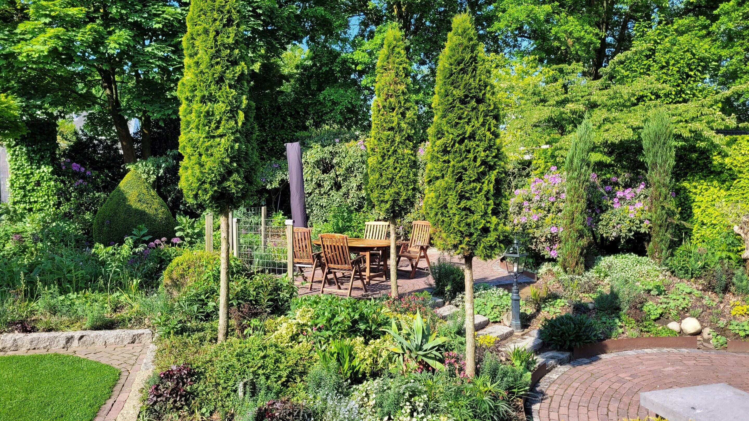 A garden with a wooden table and chairs, surrounded by various plants and trees.