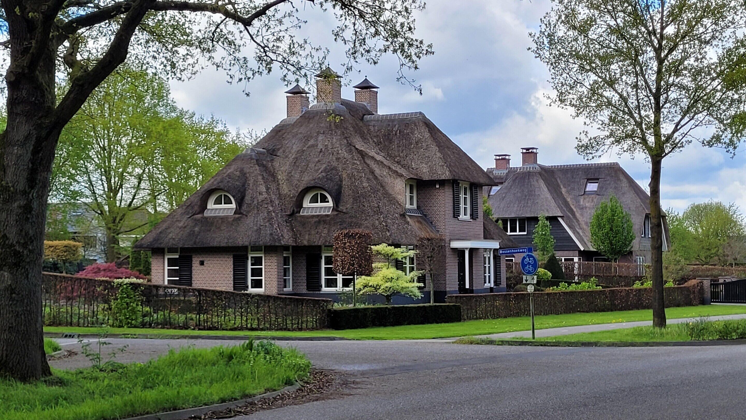Une maison traditionnelle à toit de chaume, avec une extension en briques, un panneau indiquant la présence d’un service de location de vélos et un jardin bien entretenu.
