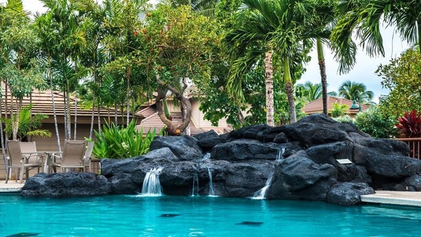 Une piscine agrémentée d’une cascade, entourée de palmiers, et un bâtiment doté d’un toit en tuiles.