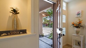 Un couloir avec une porte donnant sur l’extérieur, une petite table sur laquelle est posé un vase, et une pancarte sur laquelle est écrit le mot « aloha ».