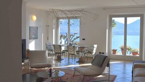 Ett rymligt vardagsrum med ett matbord, flera stolar, en soffa och ett stort fönster med utsikt över havet och bergen.