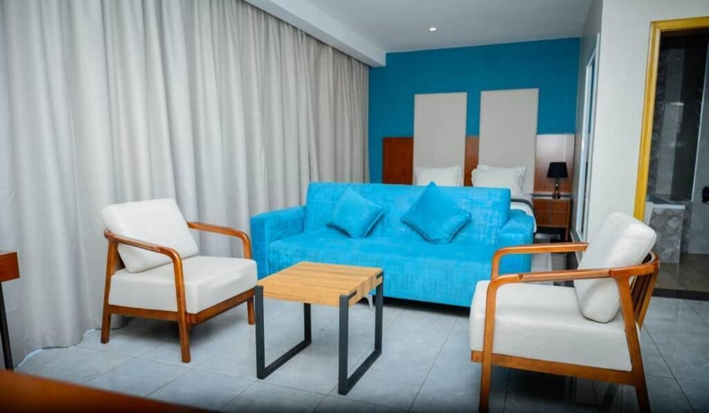 Una habitación de hotel con un sofá azul, dos sillones blancos, una mesa de centro de madera y una cama con ropa blanca.