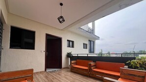 A covered outdoor seating area with wooden furniture and orange cushions, a door, and a view of the cityscape. - EDS Hébergement meublé (Abidjan)