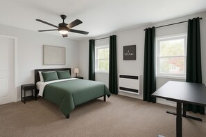 Une chambre à coucher comprenant un lit, une table de chevet, un bureau, un ventilateur de plafond, une fenêtre avec des rideaux et un radiateur fixé au mur.