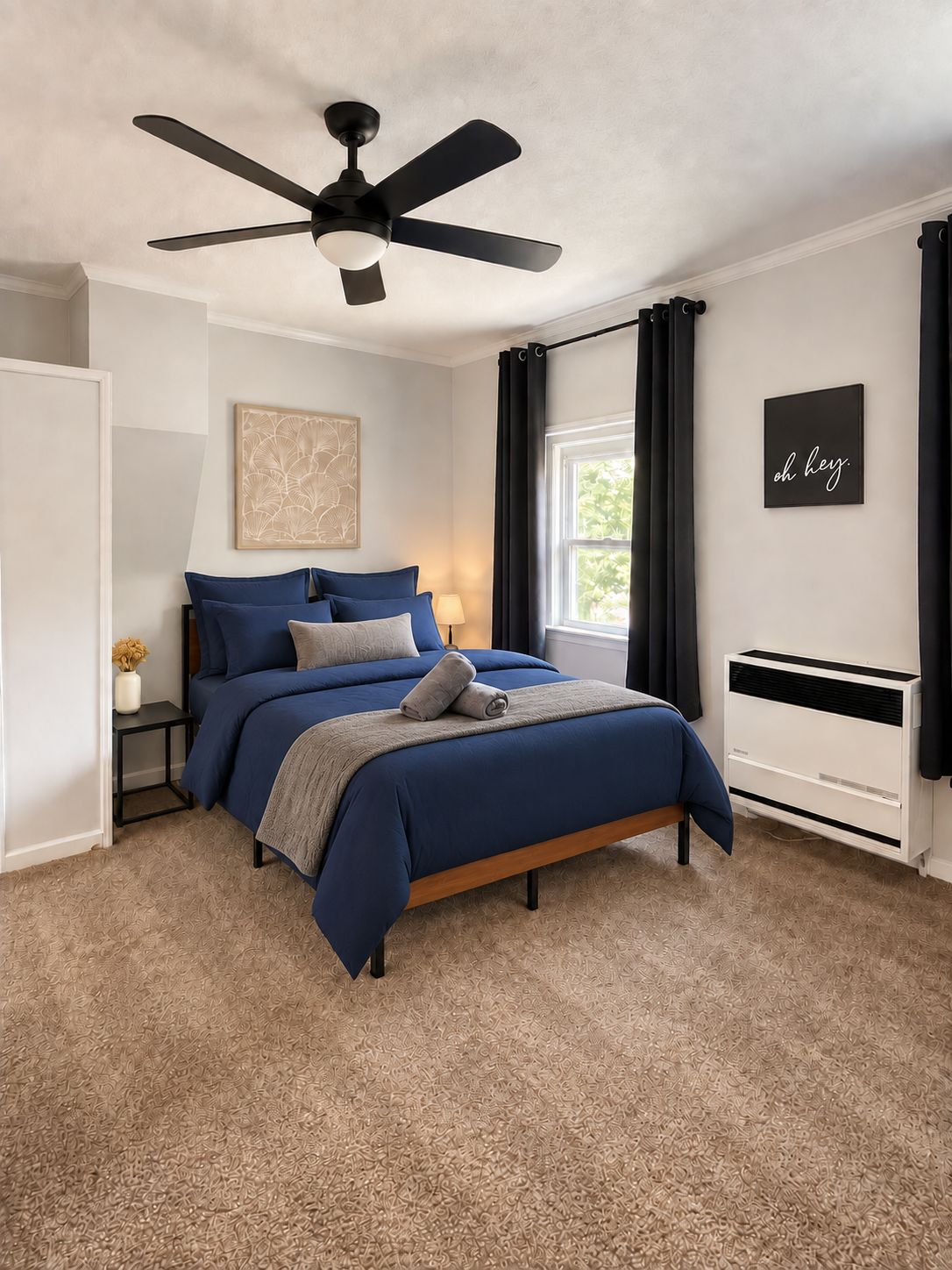 Une chambre à coucher comprenant un lit, un ventilateur de plafond, une fenêtre avec des rideaux, un climatiseur fixé au mur et une œuvre d’art encadrée.