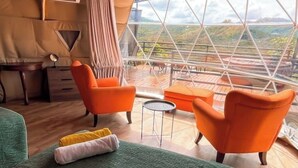 A room with a bed, two orange chairs, a small round table, and a view of a mountainous landscape through a large window. - Dome 1 Potrero Beach | Natural Getaway with Ocean View and Private Jacuzzi (Potrero)