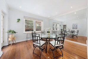 Un espace repas avec une table en verre entourée de chaises noires dotées de coussins blancs, un plancher en bois et de grandes fenêtres équipées de stores.