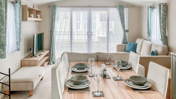 Quarto moderno e bem iluminado, com mesa de jantar posta para quatro pessoas, sofá, televisão e vista para uma varanda.