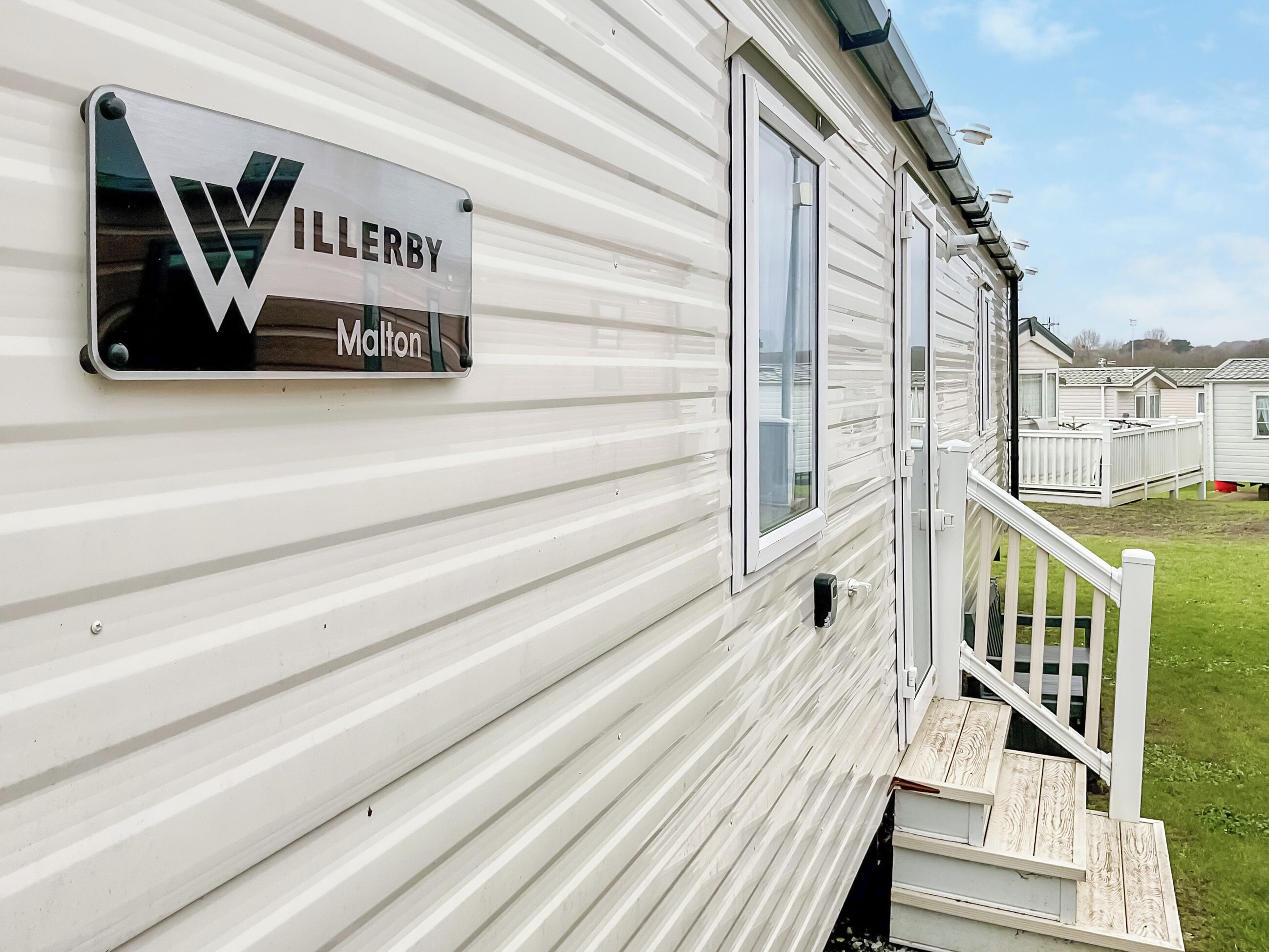 A mobile home with a sign that reads 'WILLERBY Malton' and a wooden staircase leading to a deck.