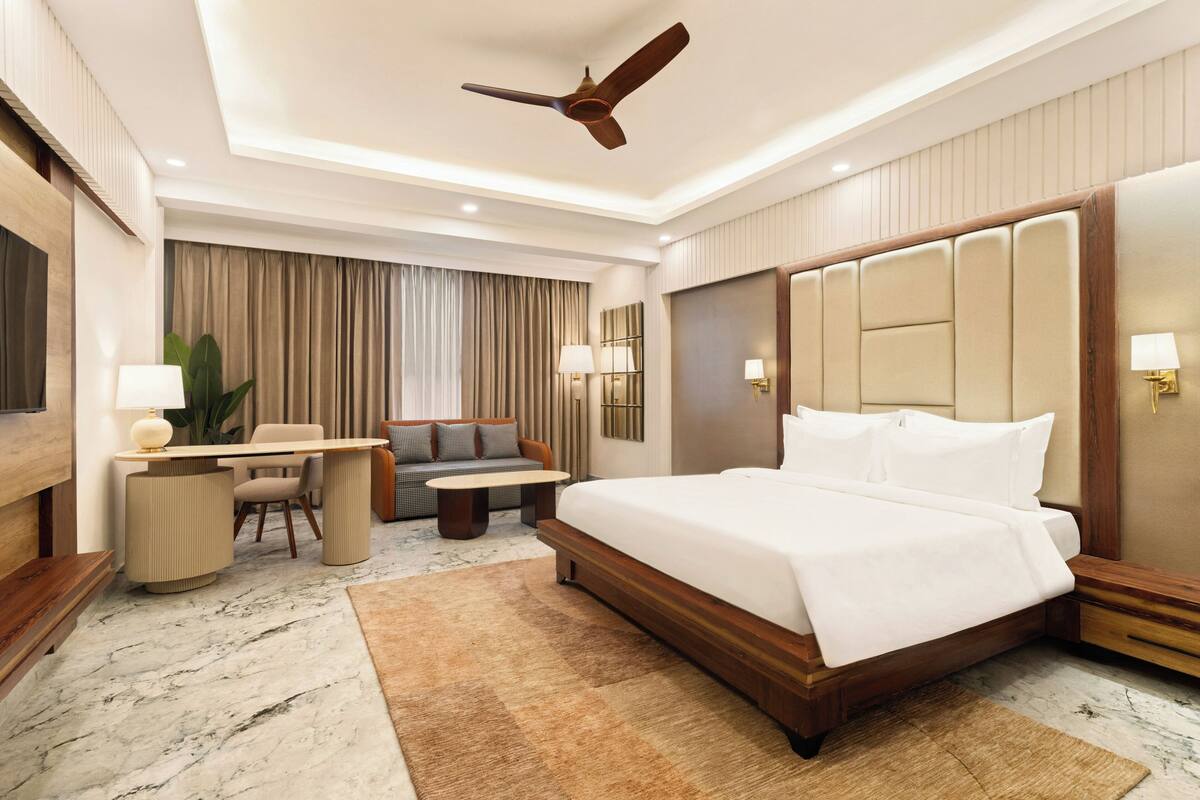 Habitación de hotel con una cama grande, un escritorio con silla, un sofá, un ventilador de techo y un televisor.