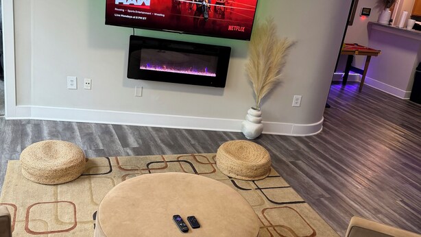 A living room with a flat-screen TV mounted on the wall, a round ottoman, and a patterned rug. - Welcome to your fun 2BR/2BA arcade loft—relax, play, and enjoy your stay!! (Atlanta)