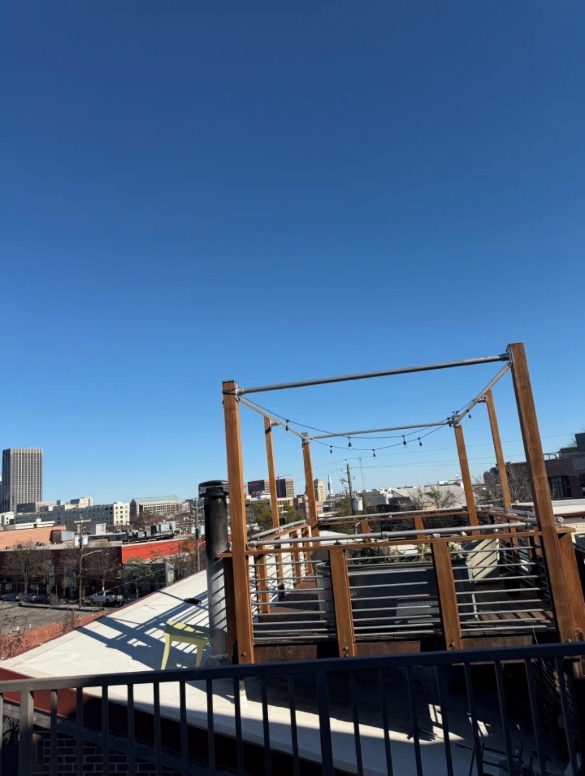 Eine Dachterrasse mit einer Holzkonstruktion, möglicherweise ein Deck oder Spielbereich, mit Blick auf eine Stadtlandschaft aus Gebäuden und einem klaren, blauen Himmel.