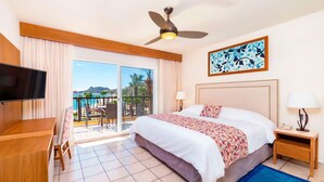 Quarto de hotel com cama, televisão, secretária, varanda com vista para o mar e ventoinha no teto.