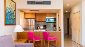 Cozinha compacta com geladeira, micro-ondas e cafeteira. Há duas cadeiras cor-de-rosa e uma mesa pequena com luminária. Um quadro está na parede.