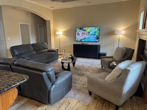 Een zithoek met een zwarte leren bank, een beige fauteuil, een salontafel en een flatscreen televisie aan de muur.