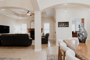 Een ruime woonkamer met een grote flatscreen-tv, een donkergrijze bank, een eettafel met stoelen en een decoratieve vaas.