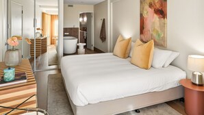 Une chambre d’hôtel moderne avec un grand lit, une table de chevet, un miroir et une salle de bain visible à travers une porte ouverte.
