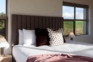 A bedroom with a bed, pillows, a nightstand, a lamp, and a window with a view of a grassy field.