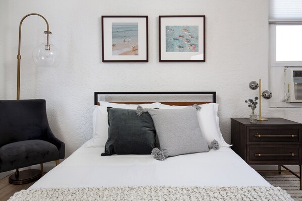 Quarto com cama, uma cadeira, mesa de cabeceira e duas quadros na parede.