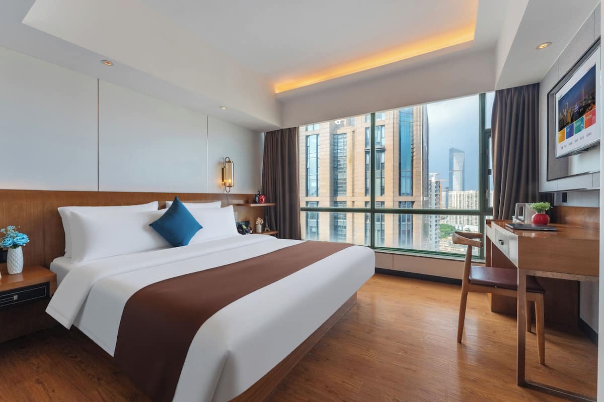 Ein Hotelzimmer mit einem großen Bett, einem Schreibtisch, einem Sessel, einem Fernseher und Blick auf hohe Gebäude.