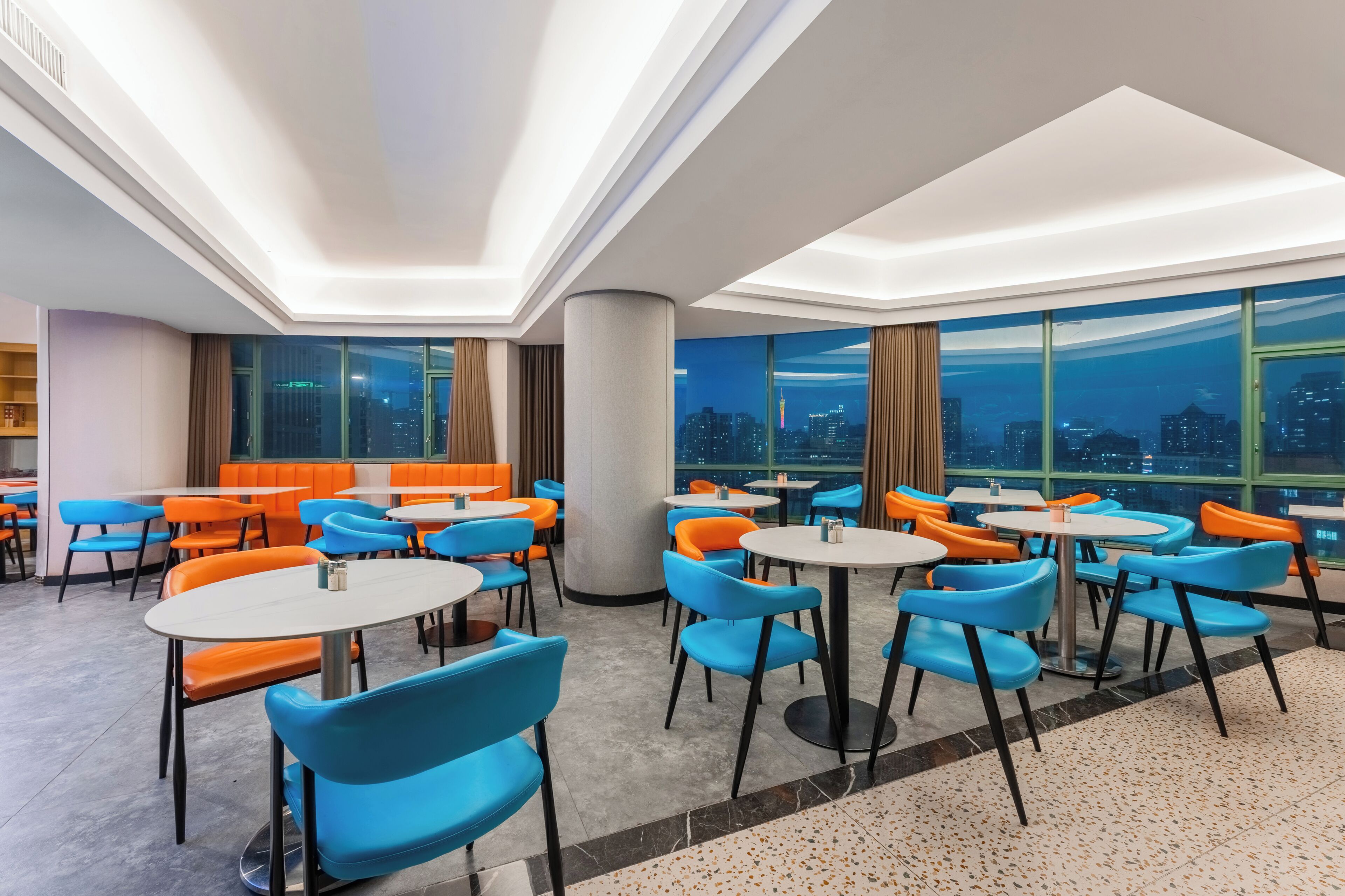 Zona pranzo moderna con tavoli e sedie rotonde, ampie finestre con vista sulla città e una colonna centrale.