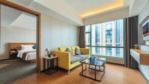 Una habitación de hotel con una cama, un sofá amarillo, una mesita de café, una mesita auxiliar pequeña y un ventanal con vistas a la ciudad.