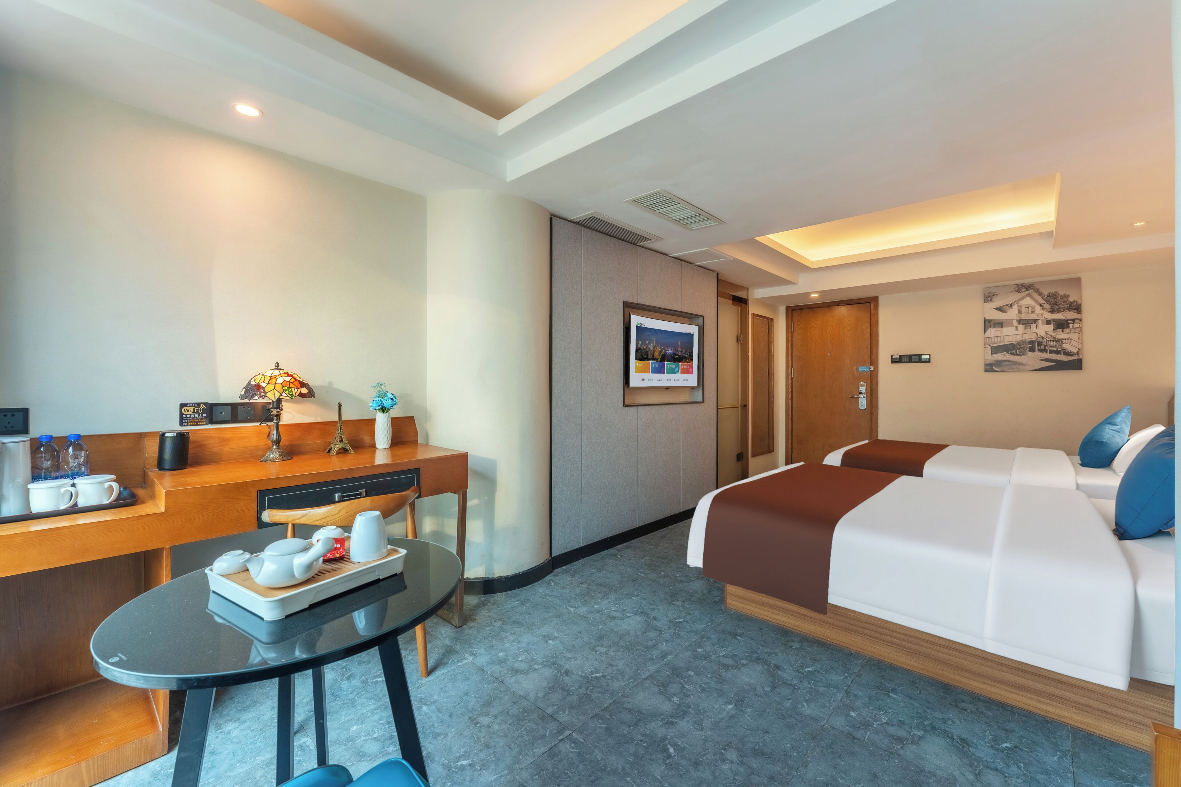 Ein Hotelzimmer mit einem Bett, einem Schreibtisch mit Stuhl, einem kleinen Tisch mit Tablett und einem an der Wand befestigten Fernseher.