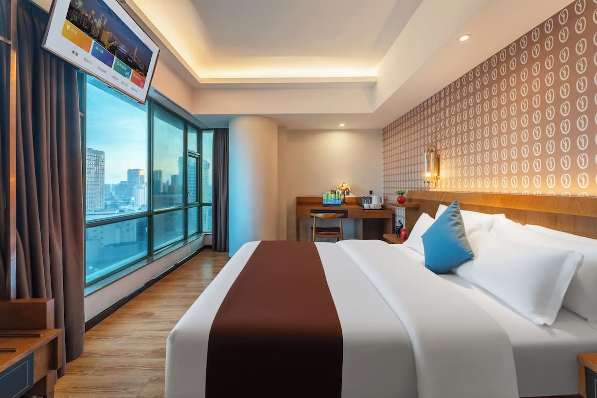 Ein Hotelzimmer mit großem Fenster, einem Bett mit weißer und brauner Bettwäsche, einem Schreibtisch mit einem Laptop und Blick auf die Stadt.