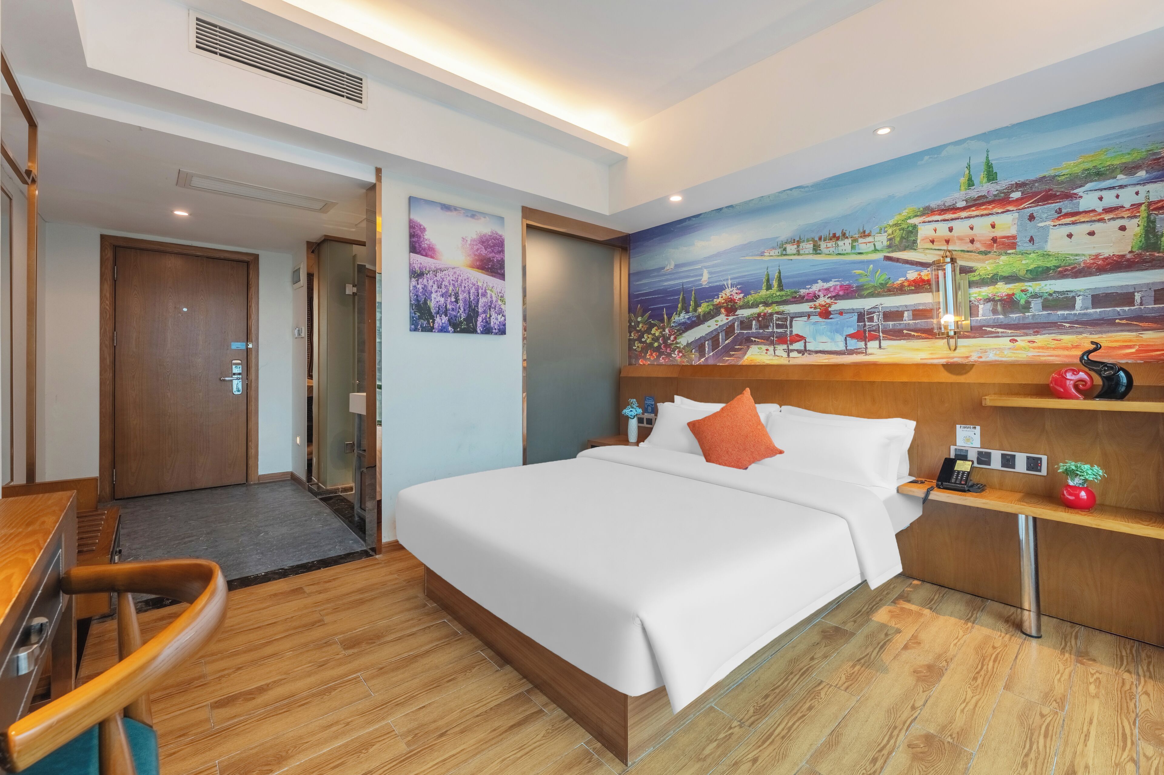 Habitación de hotel con cama, escritorio, silla, un cuadro, una lámpara y una planta pequeña.