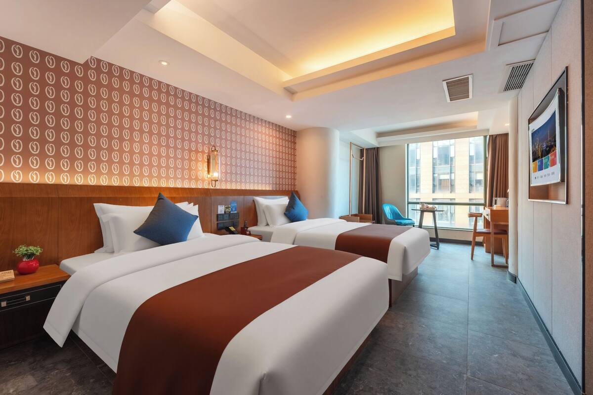Ein Hotelzimmer mit zwei Betten, einem Schreibtisch, einem Stuhl, einem kleinen Tisch, einem Fenster mit Blick und einer Wand mit Muster.