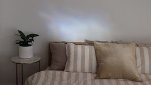 Een bed met gestreepte beddengoed, een kamerplant op een nachtkastje, en een regenboog op de achtergrond.