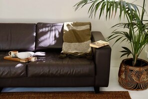 A dark brown leather sofa with a woven throw blanket and a small wooden tray with a bowl and mug. A potted plant with long green leaves is beside the sofa. - Drift Studio at Prevelly Beach (Prevelly)